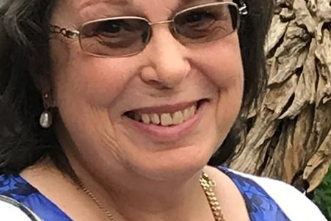 A woman with short brown hair, glasses, and a friendly smile is wearing a blue floral top, white sweater, and gold necklace. She is standing outdoors with greenery and textured bark in the background.