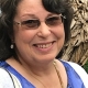 A woman with short brown hair, glasses, and a friendly smile is wearing a blue floral top, white sweater, and gold necklace. She is standing outdoors with greenery and textured bark in the background.